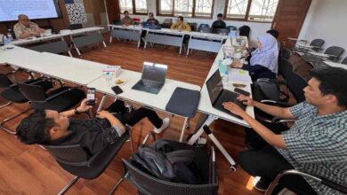 Suasana rapat revisi kurikulum PTI UMS bersama dosen dan staf di ruang rapat dengan lantai kayu. Tampak layar monitor menampilkan dokumen akademik dan beberapa peserta menggunakan laptop serta smartphone.