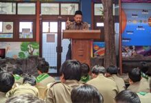 Penceramah berpeci dan berseragam coklat, berdiri di podium kayu, menyampaikan presentasi kepada siswa yang duduk di lantai di depan latar belakang poster sistem tata surya berwarna biru di SD Muhammadiyah 1 Solo.