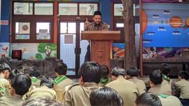 Penceramah berpeci dan berseragam coklat, berdiri di podium kayu, menyampaikan presentasi kepada siswa yang duduk di lantai di depan latar belakang poster sistem tata surya berwarna biru di SD Muhammadiyah 1 Solo.