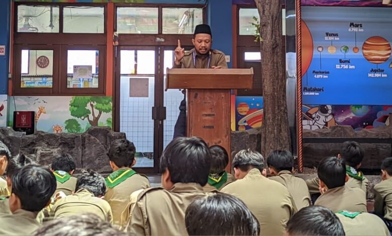 Penceramah berpeci dan berseragam coklat, berdiri di podium kayu, menyampaikan presentasi kepada siswa yang duduk di lantai di depan latar belakang poster sistem tata surya berwarna biru di SD Muhammadiyah 1 Solo.