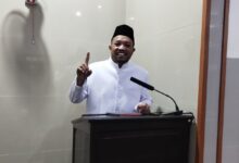 Ustaz Dwi Jatmiko, Dai MUI Pusat, menyampaikan tausiyah di Masjid Al Mukarromah Solo tentang lima fase kehidupan manusia dan bekal akhirat. Dia berdiri di belakang podium sambil tersenyum dan mengangkat jari telunjuk, dengan latar belakang dinding keramik abu-abu muda dan kipas angin gantung.