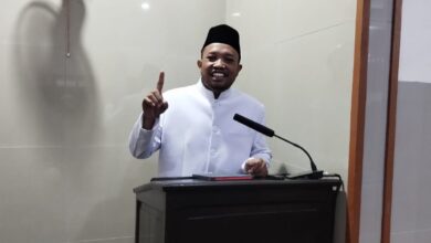 Ustaz Dwi Jatmiko, Dai MUI Pusat, menyampaikan tausiyah di Masjid Al Mukarromah Solo tentang lima fase kehidupan manusia dan bekal akhirat. Dia berdiri di belakang podium sambil tersenyum dan mengangkat jari telunjuk, dengan latar belakang dinding keramik abu-abu muda dan kipas angin gantung.