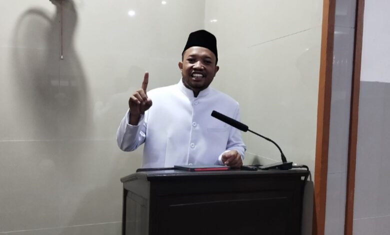 Ustaz Dwi Jatmiko, Dai MUI Pusat, menyampaikan tausiyah di Masjid Al Mukarromah Solo tentang lima fase kehidupan manusia dan bekal akhirat. Dia berdiri di belakang podium sambil tersenyum dan mengangkat jari telunjuk, dengan latar belakang dinding keramik abu-abu muda dan kipas angin gantung.
