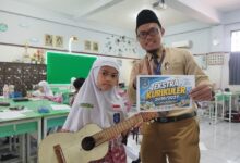 Siswi memegang gitar ukulele dan guru menunjukkan poster promosi ekstrakurikuler SD Muhammadiyah 1 Solo di dalam ruang kelas.