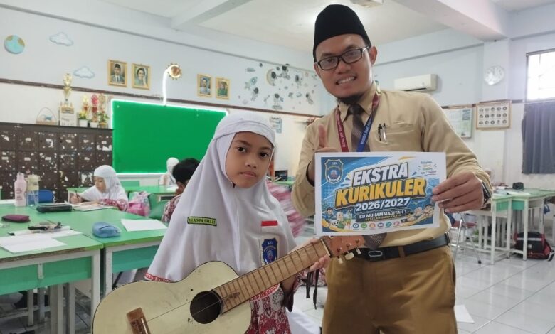 Siswi memegang gitar ukulele dan guru menunjukkan poster promosi ekstrakurikuler SD Muhammadiyah 1 Solo di dalam ruang kelas.