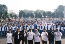 Ribuan pelajar Muhammadiyah Wonosobo mengenakan seragam biru-putih dan jilbab memadati Halaman Sasana Adipura, berpose semangat dengan kepalan tangan. Di latar depan, tamu VIP mengenakan kaus acara berlogo SMUFest 2026 Wonosobo. Latar belakang menampilkan Alun-Alun Wonosobo. Gambar mendokumentasikan puncak Fortasi 2026.