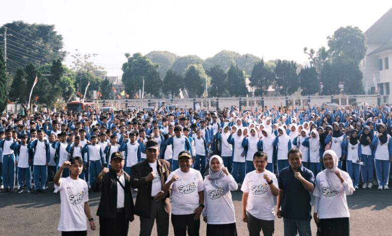 Ribuan pelajar Muhammadiyah Wonosobo mengenakan seragam biru-putih dan jilbab memadati Halaman Sasana Adipura, berpose semangat dengan kepalan tangan. Di latar depan, tamu VIP mengenakan kaus acara berlogo SMUFest 2026 Wonosobo. Latar belakang menampilkan Alun-Alun Wonosobo. Gambar mendokumentasikan puncak Fortasi 2026.