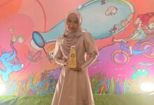 Seorang wanita muda berhijab, diidentifikasi sebagai Khanza Kineta Asmarani dari SMP Mutual Magelang, tersenyum bangga sambil memegang piala penghargaan juara menyanyi. Dia mengenakan gaun merah muda keunguan dengan latar belakang panggung bertema kartun anak yang penuh warna.