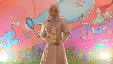 Seorang wanita muda berhijab, diidentifikasi sebagai Khanza Kineta Asmarani dari SMP Mutual Magelang, tersenyum bangga sambil memegang piala penghargaan juara menyanyi. Dia mengenakan gaun merah muda keunguan dengan latar belakang panggung bertema kartun anak yang penuh warna.
