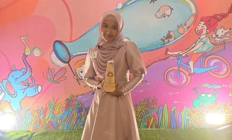 Seorang wanita muda berhijab, diidentifikasi sebagai Khanza Kineta Asmarani dari SMP Mutual Magelang, tersenyum bangga sambil memegang piala penghargaan juara menyanyi. Dia mengenakan gaun merah muda keunguan dengan latar belakang panggung bertema kartun anak yang penuh warna.