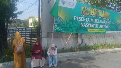 Tiga peserta wanita Muktamar Nasyiatul Aisyiyah Solo sedang beristirahat di pinggir jalan di depan spanduk ucapan selamat datang besar berwarna hijau. Spanduk tersebut memuat teks "Selamat Datang Peserta Muktamar ke-15 Nasyiatul Aisyiyah" dan tanggal acara di Surakarta, 6-8 Agustus 2026.