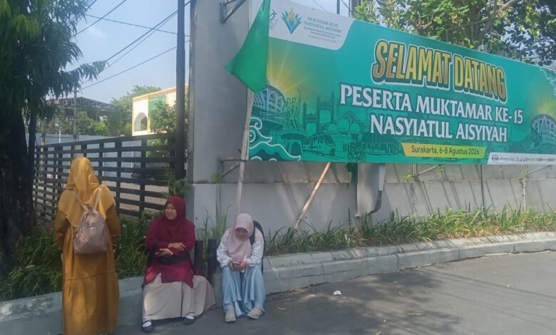 Tiga peserta wanita Muktamar Nasyiatul Aisyiyah Solo sedang beristirahat di pinggir jalan di depan spanduk ucapan selamat datang besar berwarna hijau. Spanduk tersebut memuat teks "Selamat Datang Peserta Muktamar ke-15 Nasyiatul Aisyiyah" dan tanggal acara di Surakarta, 6-8 Agustus 2026.