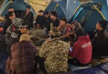 Sekelompok pria duduk melingkar di atas alas biru sambil menikmati makan malam bersama di tengah perkemahan di Embung Kledung, dengan latar belakang tenda-tenda berwarna biru dan pink pada malam hari.