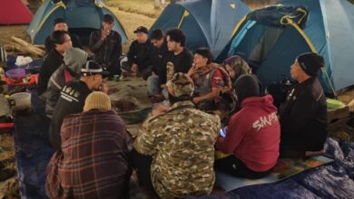 Sekelompok pria duduk melingkar di atas alas biru sambil menikmati makan malam bersama di tengah perkemahan di Embung Kledung, dengan latar belakang tenda-tenda berwarna biru dan pink pada malam hari.