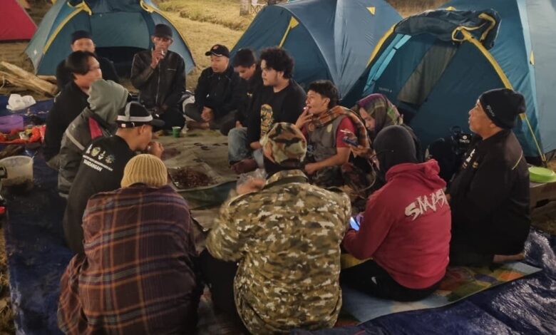 Sekelompok pria duduk melingkar di atas alas biru sambil menikmati makan malam bersama di tengah perkemahan di Embung Kledung, dengan latar belakang tenda-tenda berwarna biru dan pink pada malam hari.
