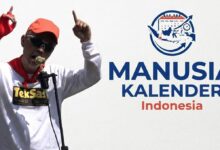 Zenza TekSas sang Manusia Kalender Indonesia tampil mengenakan topi oranye dan kacamata hitam sambil menunjuk ke atas di depan mikrofon.