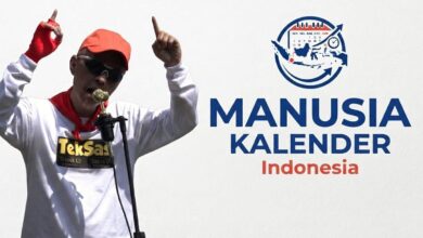 Zenza TekSas sang Manusia Kalender Indonesia tampil mengenakan topi oranye dan kacamata hitam sambil menunjuk ke atas di depan mikrofon.