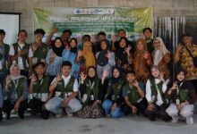 Foto bersama tim mahasiswa PPK Ormawa Unimus dan warga Pringapus dalam kegiatan pelatihan pemasaran digital produk, berpose membentuk tanda 'OK' di depan spanduk acara di dalam ruangan.