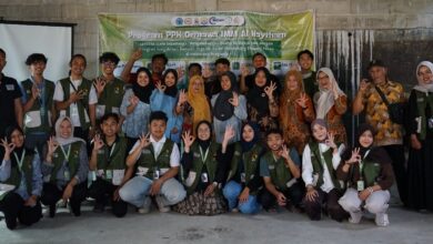 Foto bersama tim mahasiswa PPK Ormawa Unimus dan warga Pringapus dalam kegiatan pelatihan pemasaran digital produk, berpose membentuk tanda 'OK' di depan spanduk acara di dalam ruangan.