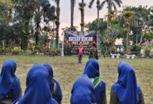 Suasana apel pembukaan Diksar KOKAM Pelajar Wonosobo dengan peserta berhijab biru berbaris rapi menghadap instruktur dan spanduk acara di area outdoor yang asri di The Lodjie Marong, Wonosobo.