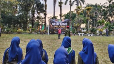 Suasana apel pembukaan Diksar KOKAM Pelajar Wonosobo dengan peserta berhijab biru berbaris rapi menghadap instruktur dan spanduk acara di area outdoor yang asri di The Lodjie Marong, Wonosobo.