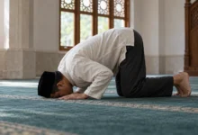 Seorang pria berpeci hitam bersujud di atas karpet biru dalam masjid yang diterangi cahaya alami dari jendela.