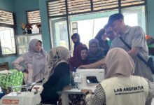 Sekelompok orang sedang mengerumuni sebuah mesin jahit di dalam sebuah ruangan bengkel, mendengarkan penjelasan dari seorang instruktur tentang cara mengoperasikannya dalam rangka pelatihan produksi batik ciprat.