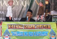 Seorang pria berkacamata dan berpeci berbicara di mikrofon di belakang meja yang bertuliskan "Wayang Golek Pitutur 'MARSUDI ING REH KAUTAMEN'", diapit oleh dua pasang wayang golek berbusana adat Jawa.