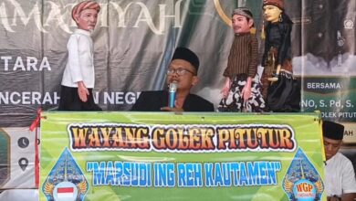 Seorang pria berkacamata dan berpeci berbicara di mikrofon di belakang meja yang bertuliskan "Wayang Golek Pitutur 'MARSUDI ING REH KAUTAMEN'", diapit oleh dua pasang wayang golek berbusana adat Jawa.