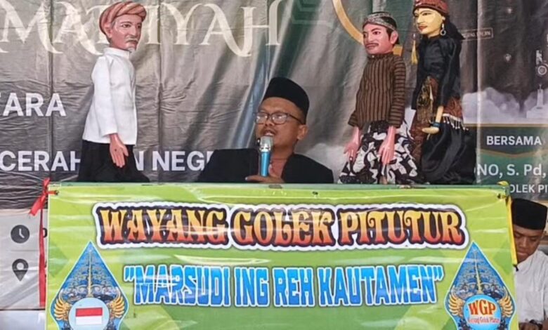 Seorang pria berkacamata dan berpeci berbicara di mikrofon di belakang meja yang bertuliskan "Wayang Golek Pitutur 'MARSUDI ING REH KAUTAMEN'", diapit oleh dua pasang wayang golek berbusana adat Jawa.