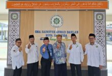 Enam pria dalam balutan seragam berpose 'finger heart' di depan lobi SMA Sainstek Ahmad Dahlan Salatiga, di bawah kaligrafi surah Ali Imran ayat 104, melambangkan semangat sinergi dan optimisme FKKM Karanggede dan FGM Pusat.