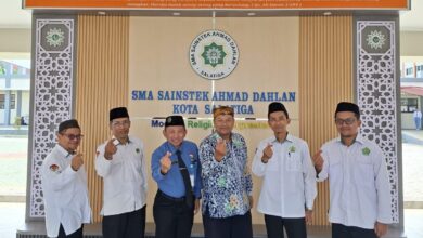 Enam pria dalam balutan seragam berpose 'finger heart' di depan lobi SMA Sainstek Ahmad Dahlan Salatiga, di bawah kaligrafi surah Ali Imran ayat 104, melambangkan semangat sinergi dan optimisme FKKM Karanggede dan FGM Pusat.
