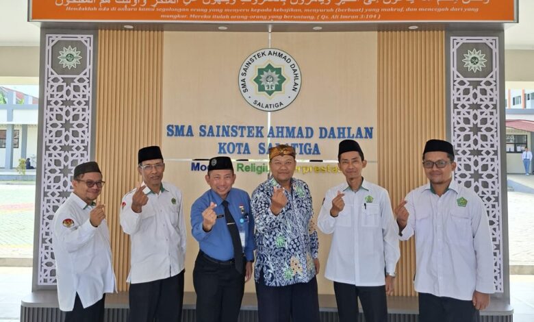Enam pria dalam balutan seragam berpose 'finger heart' di depan lobi SMA Sainstek Ahmad Dahlan Salatiga, di bawah kaligrafi surah Ali Imran ayat 104, melambangkan semangat sinergi dan optimisme FKKM Karanggede dan FGM Pusat.