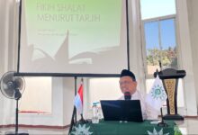 Prof Ahwan Fanani duduk di meja berlogo Muhammadiyah memberikan materi Fikih Shalat Tarjih Muhammadiyah dengan latar belakang layar proyektor.