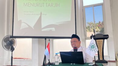 Prof Ahwan Fanani duduk di meja berlogo Muhammadiyah memberikan materi Fikih Shalat Tarjih Muhammadiyah dengan latar belakang layar proyektor.