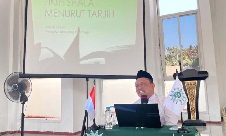Prof Ahwan Fanani duduk di meja berlogo Muhammadiyah memberikan materi Fikih Shalat Tarjih Muhammadiyah dengan latar belakang layar proyektor.