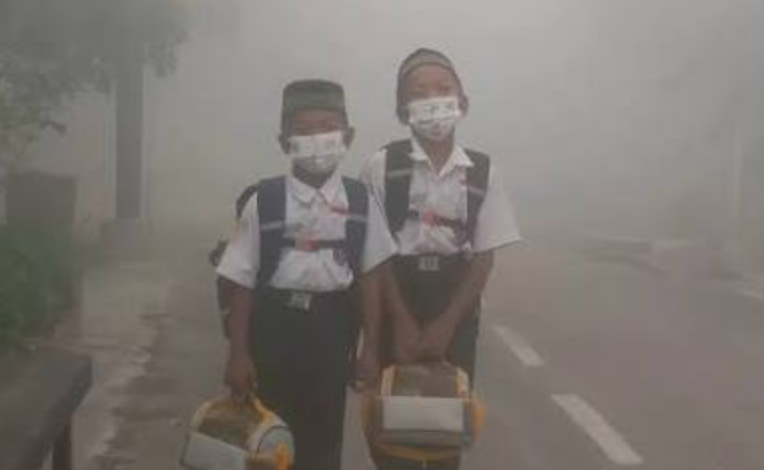 Dua anak sekolah dasar memakai masker medis di tengah kepungan kabut asap tebal akibat dampak karhutla pada anak.
